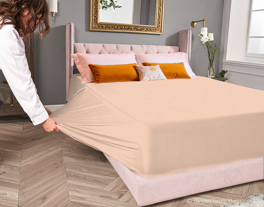 Apricot jersey-knit stretch fitted sheet on an extra-deep mattress — pulling the sidewall over a foot outward to demonstrate the abundant stretch, making sheet changing effortless even for those with limited strength or mobility.
