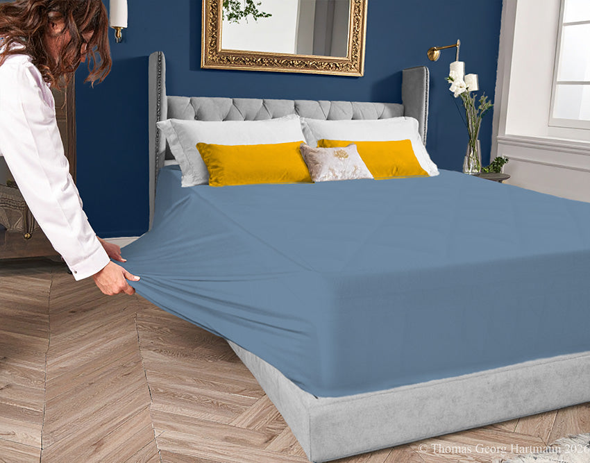 Azure jersey-knit stretch fitted sheet on an extra-deep mattress — pulling the sidewall over a foot outward to demonstrate the abundant stretch, making sheet changing effortless even for those with limited strength or mobility.