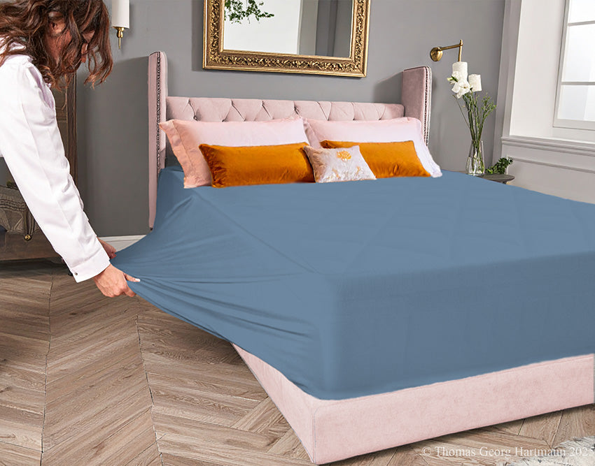 Azure jersey-knit stretch fitted sheet on an extra-deep mattress — pulling the sidewall over a foot outward to demonstrate the abundant stretch, making sheet changing effortless even for those with limited strength or mobility.
