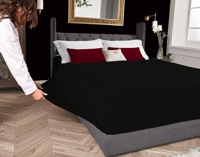Black jersey-knit stretch fitted sheet on an extra-deep mattress — pulling the sidewall over a foot outward to demonstrate the abundant stretch, making sheet changing effortless even for those with limited strength or mobility.
