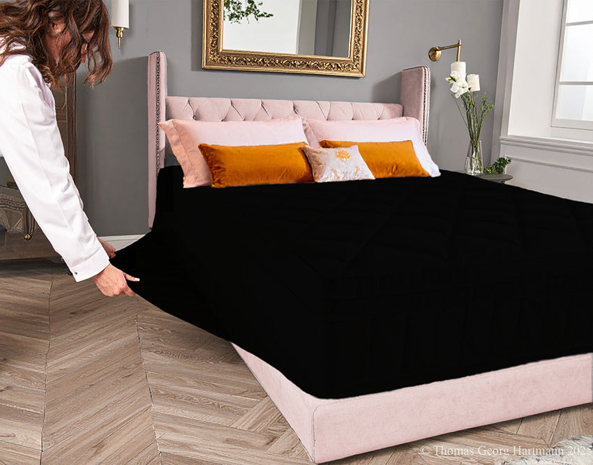 Black jersey-knit stretch fitted sheet on an extra-deep mattress — pulling the sidewall over a foot outward to demonstrate the abundant stretch, making sheet changing effortless even for those with limited strength or mobility.