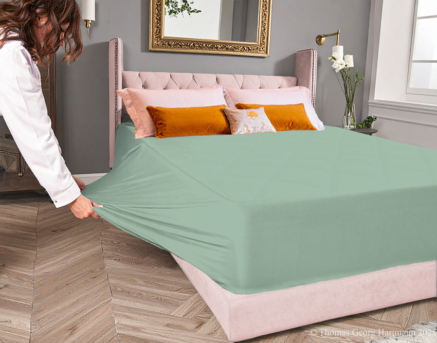 Blue-grass jersey-knit stretch fitted sheet on an extra-deep mattress — pulling the sidewall over a foot outward to demonstrate the abundant stretch, making sheet changing effortless even for those with limited strength or mobility.