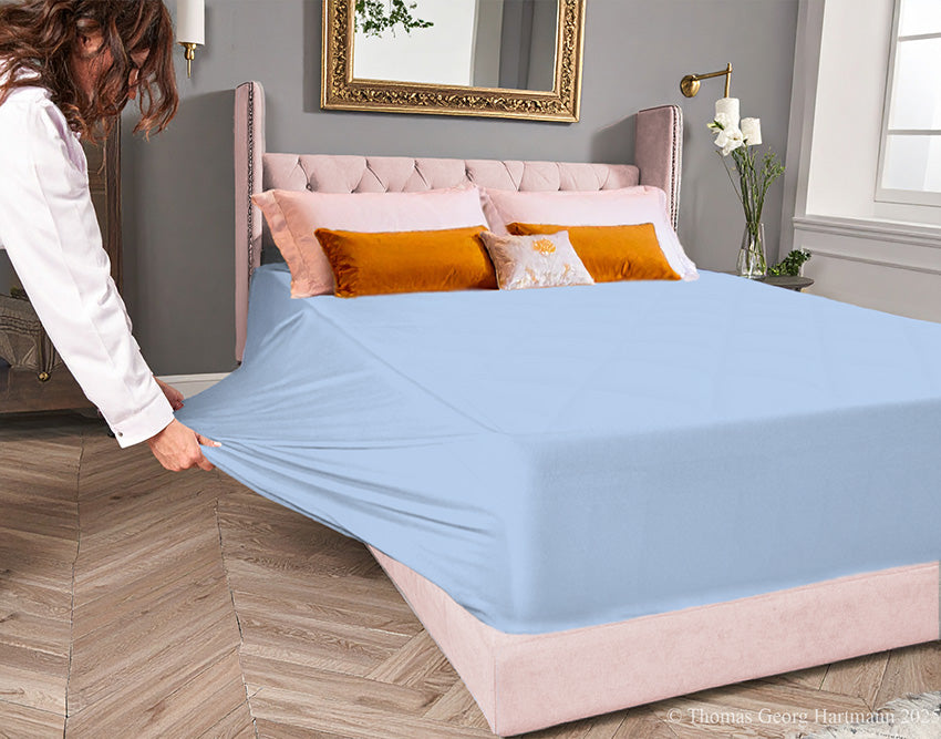 Bleu jersey-knit stretch fitted sheet on an extra-deep mattress — pulling the sidewall over a foot outward to demonstrate the abundant stretch, making sheet changing effortless even for those with limited strength or mobility.