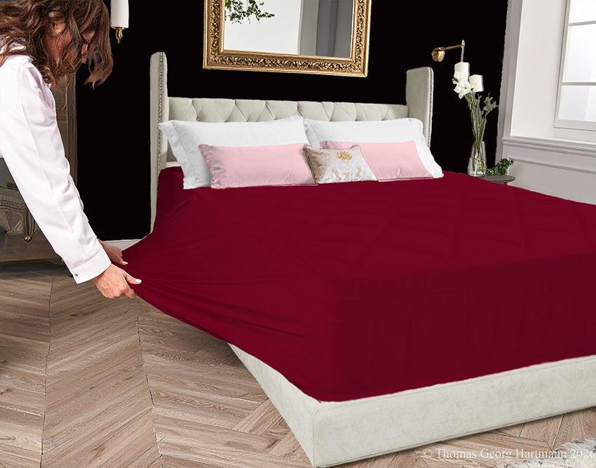 Bordeaux jersey-knit stretch fitted sheet on an extra-deep mattress — pulling the sidewall over a foot outward to demonstrate the abundant stretch, making sheet changing effortless even for those with limited strength or mobility.