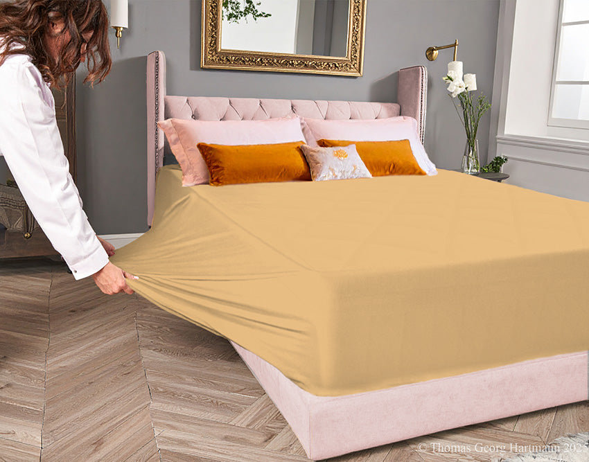 Caramel jersey-knit stretch fitted sheet on an extra-deep mattress — pulling the sidewall over a foot outward to demonstrate the abundant stretch, making sheet changing effortless even for those with limited strength or mobility.