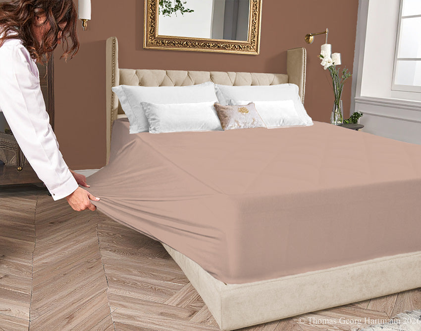 Chocolate-milk jersey-knit stretch fitted sheet on an extra-deep mattress — pulling the sidewall over a foot outward to demonstrate the abundant stretch, making sheet changing effortless even for those with limited strength or mobility.