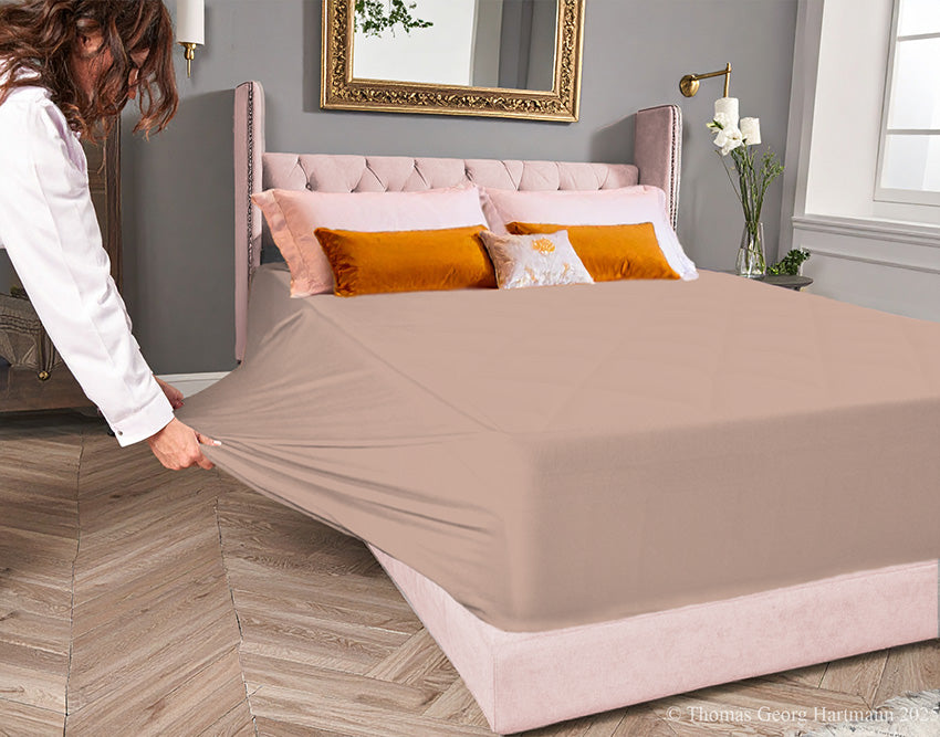 Chocolate-milk jersey-knit stretch fitted sheet on an extra-deep mattress — pulling the sidewall over a foot outward to demonstrate the abundant stretch, making sheet changing effortless even for those with limited strength or mobility.