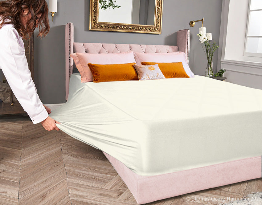 Cream jersey-knit stretch fitted sheet on an extra-deep mattress — pulling the sidewall over a foot outward to demonstrate the abundant stretch, making sheet changing effortless even for those with limited strength or mobility.