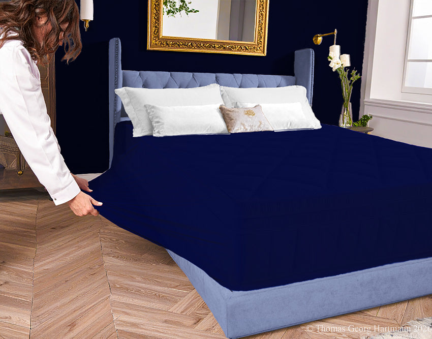 Dark-navy jersey-knit stretch fitted sheet on an extra-deep mattress — pulling the sidewall over a foot outward to demonstrate the abundant stretch, making sheet changing effortless even for those with limited strength or mobility.