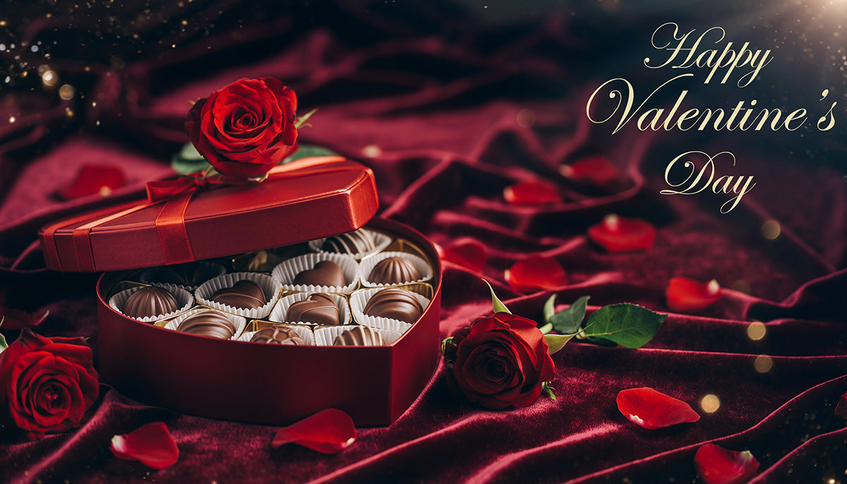 Heart-shaped box of assorted chocolates with a red rose on top, surrounded by red rose petals on dark red velvet fabric. Gold script text reads "Happy Valentine's Day" in the background.