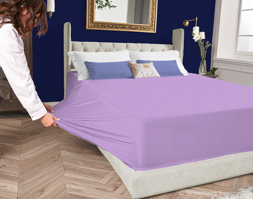 Lilac jersey-knit stretch fitted sheet on an extra-deep mattress — pulling the sidewall over a foot outward to demonstrate the abundant stretch, making sheet changing effortless even for those with limited strength or mobility.