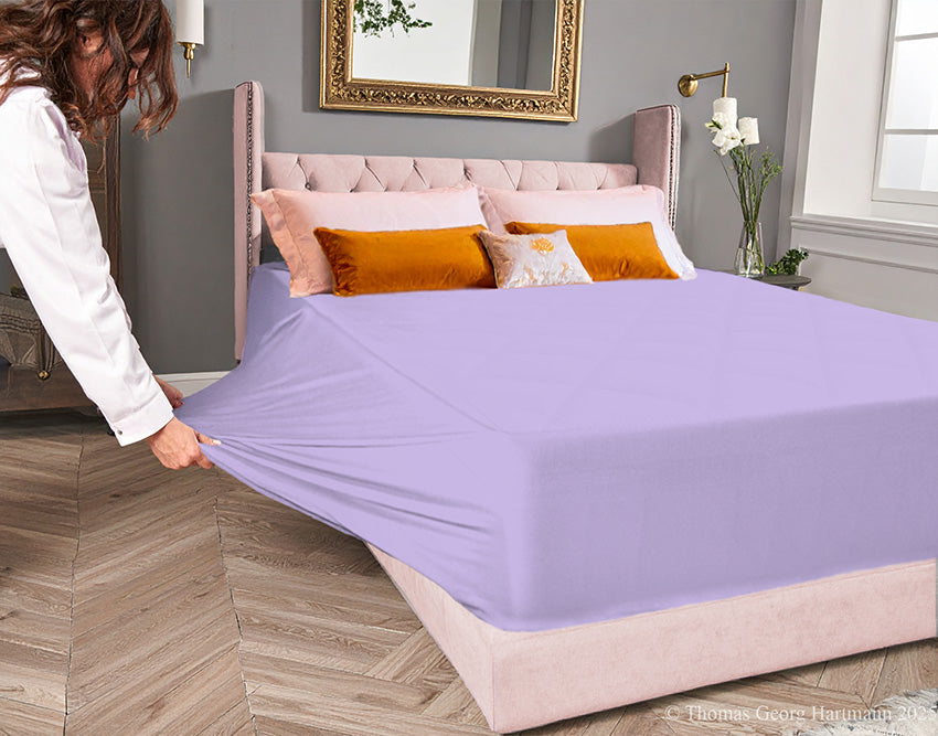 Lilac jersey-knit stretch fitted sheet on an extra-deep mattress — pulling the sidewall over a foot outward to demonstrate the abundant stretch, making sheet changing effortless even for those with limited strength or mobility.