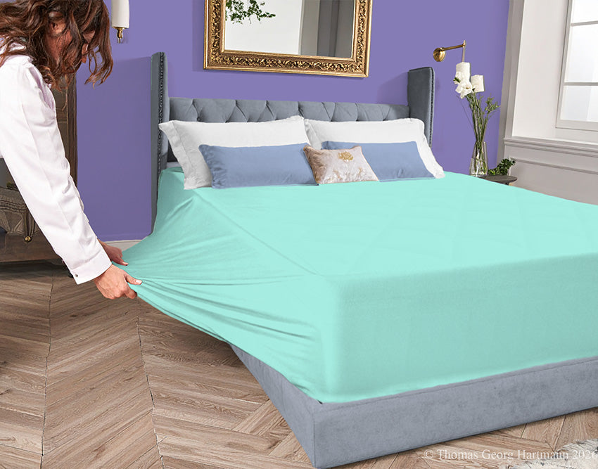 Mint jersey-knit stretch fitted sheet on an extra-deep mattress — pulling the sidewall over a foot outward to demonstrate the abundant stretch, making sheet changing effortless even for those with limited strength or mobility.
