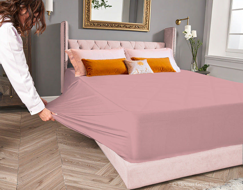 Old-rose jersey-knit stretch fitted sheet on an extra-deep mattress — pulling the sidewall over a foot outward to demonstrate the abundant stretch, making sheet changing effortless even for those with limited strength or mobility.