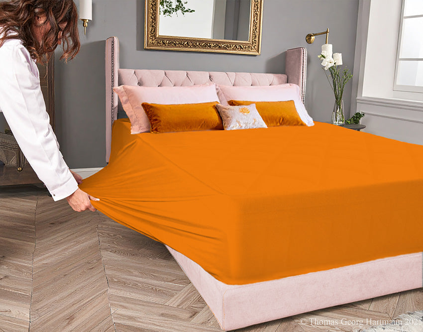 Orange jersey-knit stretch fitted sheet on an extra-deep mattress — pulling the sidewall over a foot outward to demonstrate the abundant stretch, making sheet changing effortless even for those with limited strength or mobility.