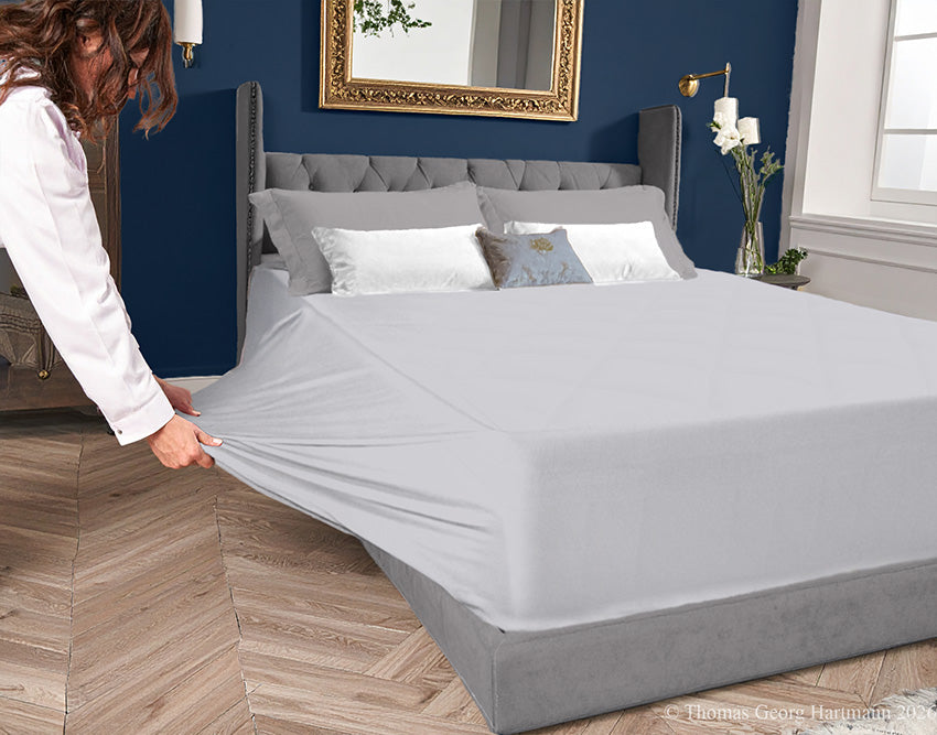 Silver jersey-knit stretch fitted sheet on an extra-deep mattress — pulling the sidewall over a foot outward to demonstrate the abundant stretch, making sheet changing effortless even for those with limited strength or mobility.