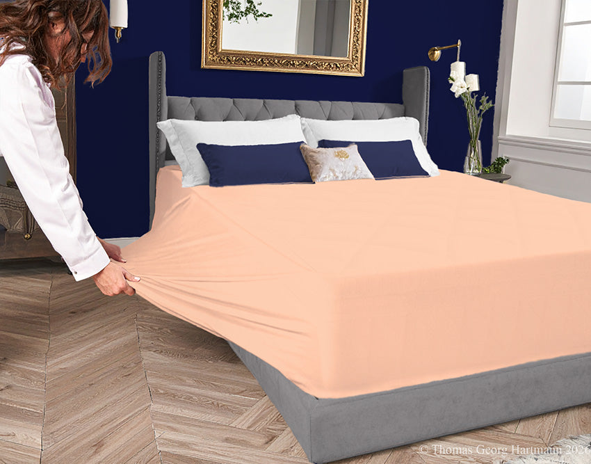Apricot jersey-knit stretch fitted sheet on an extra-deep mattress — pulling the sidewall over a foot outward to demonstrate the abundant stretch, making sheet changing effortless even for those with limited strength or mobility.