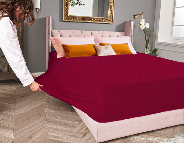 Bordeaux jersey-knit stretch fitted sheet on an extra-deep mattress — pulling the sidewall over a foot outward to demonstrate the abundant stretch, making sheet changing effortless even for those with limited strength or mobility.