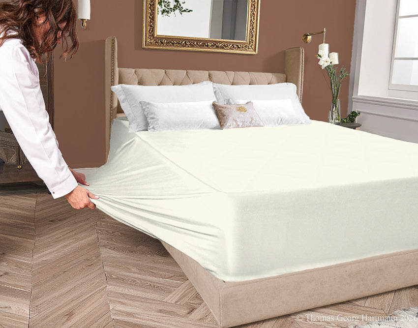 Cream jersey-knit stretch fitted sheet on an extra-deep mattress — pulling the sidewall over a foot outward to demonstrate the abundant stretch, making sheet changing effortless even for those with limited strength or mobility.