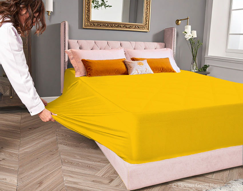 Gold jersey-knit stretch fitted sheet on an extra-deep mattress — pulling the sidewall over a foot outward to demonstrate the abundant stretch, making sheet changing effortless even for those with limited strength or mobility.
