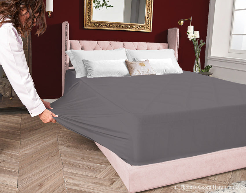 Graphite jersey-knit stretch fitted sheet on an extra-deep mattress — pulling the sidewall over a foot outward to demonstrate the abundant stretch, making sheet changing effortless even for those with limited strength or mobility.