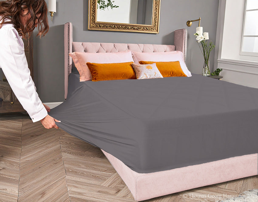 Graphite jersey-knit stretch fitted sheet on an extra-deep mattress — pulling the sidewall over a foot outward to demonstrate the abundant stretch, making sheet changing effortless even for those with limited strength or mobility.