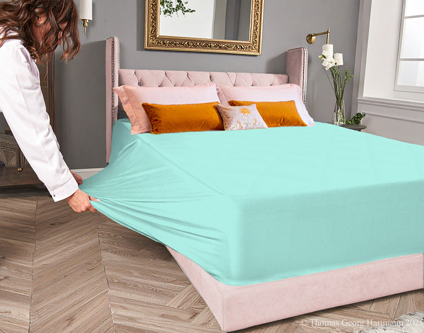 Mint jersey-knit stretch fitted sheet on an extra-deep mattress — pulling the sidewall over a foot outward to demonstrate the abundant stretch, making sheet changing effortless even for those with limited strength or mobility.