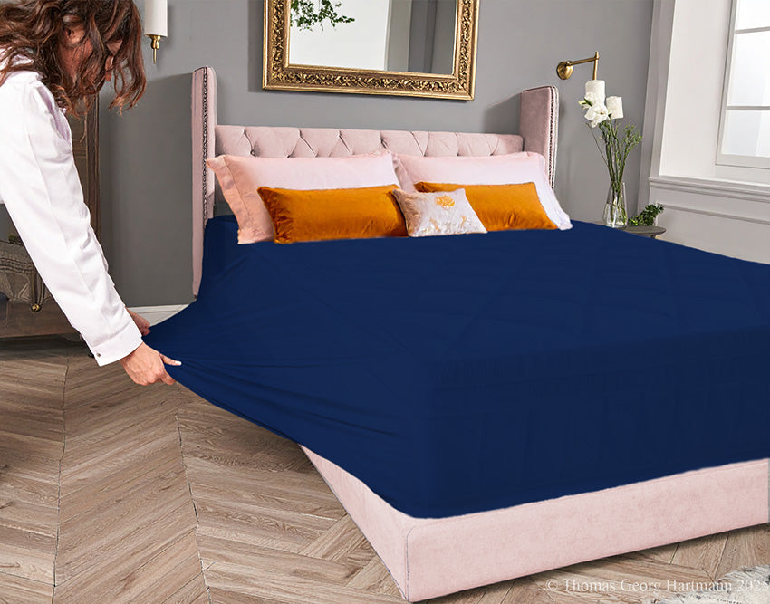 Dark-navy jersey-knit stretch fitted sheet on an extra-deep mattress — pulling the sidewall over a foot outward to demonstrate the abundant stretch, making sheet changing effortless even for those with limited strength or mobility.