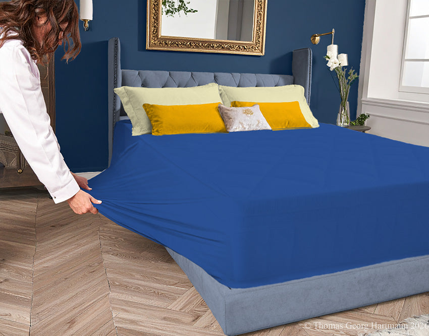 Ocean-blue jersey-knit stretch fitted sheet on an extra-deep mattress — pulling the sidewall over a foot outward to demonstrate the abundant stretch, making sheet changing effortless even for those with limited strength or mobility.