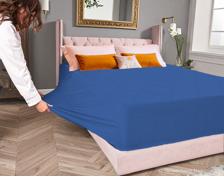 Ocean-blue jersey-knit stretch fitted sheet on an extra-deep mattress — pulling the sidewall over a foot outward to demonstrate the abundant stretch, making sheet changing effortless even for those with limited strength or mobility.