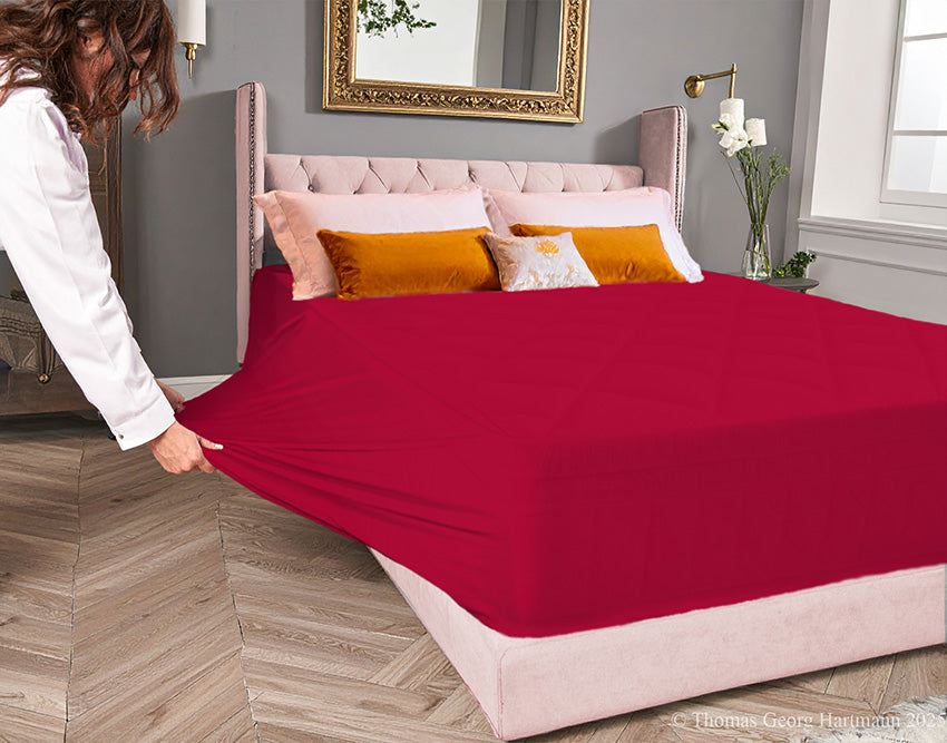 Red jersey-knit stretch fitted sheet on an extra-deep mattress — pulling the sidewall over a foot outward to demonstrate the abundant stretch, making sheet changing effortless even for those with limited strength or mobility.