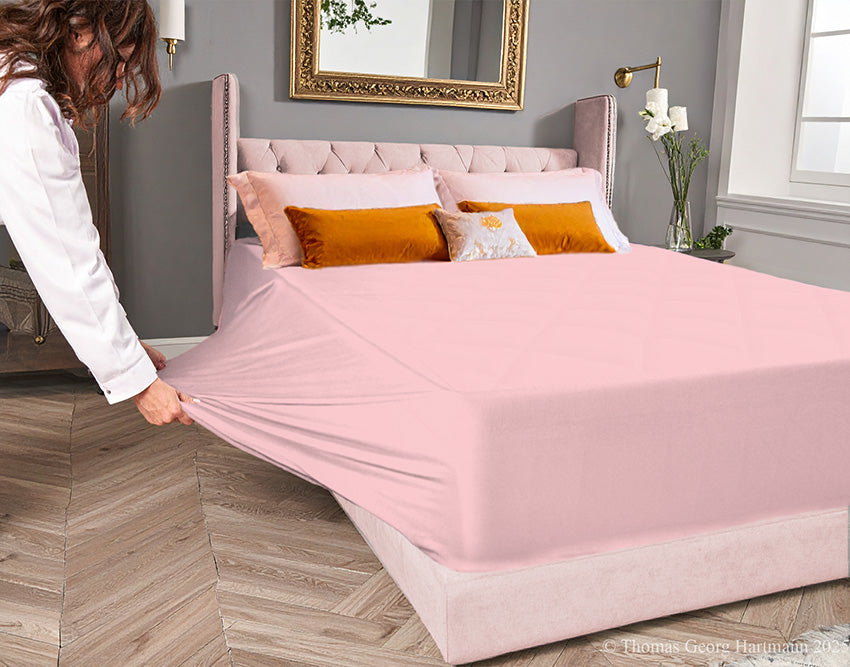Rosé jersey-knit stretch fitted sheet on an extra-deep mattress — pulling the sidewall over a foot outward to demonstrate the abundant stretch, making sheet changing effortless even for those with limited strength or mobility.