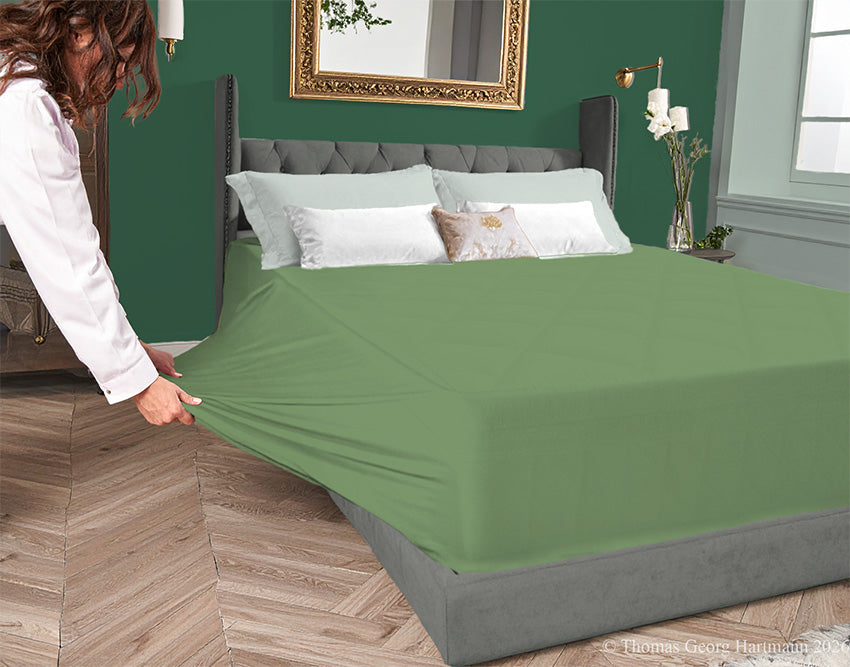 Sage jersey-knit stretch fitted sheet on an extra-deep mattress — pulling the sidewall over a foot outward to demonstrate the abundant stretch, making sheet changing effortless even for those with limited strength or mobility.