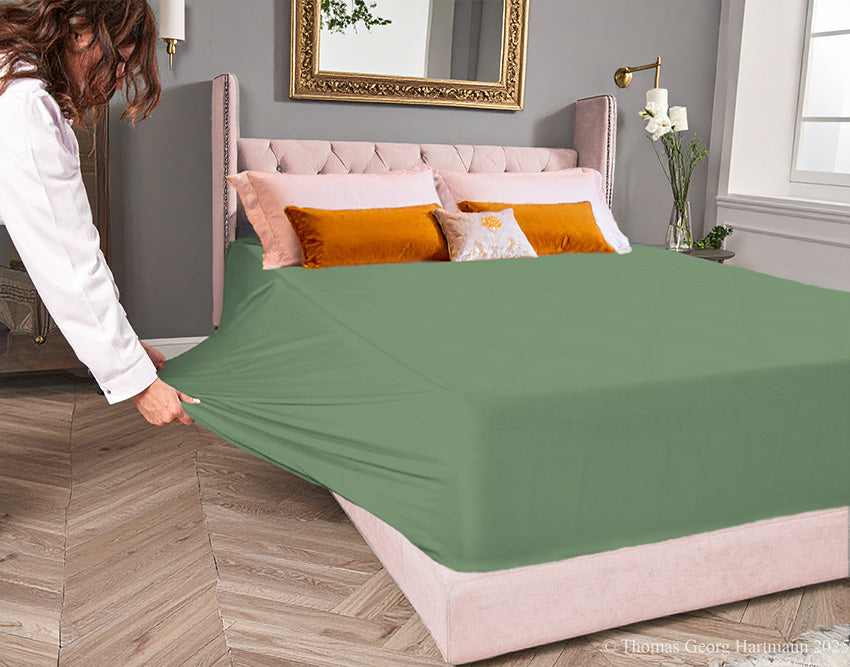 Sage jersey-knit stretch fitted sheet on an extra-deep mattress — pulling the sidewall over a foot outward to demonstrate the abundant stretch, making sheet changing effortless even for those with limited strength or mobility.