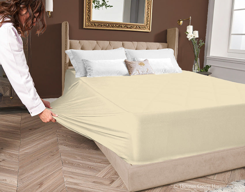 Sand jersey-knit stretch fitted sheet on an extra-deep mattress — pulling the sidewall over a foot outward to demonstrate the abundant stretch, making sheet changing effortless even for those with limited strength or mobility.