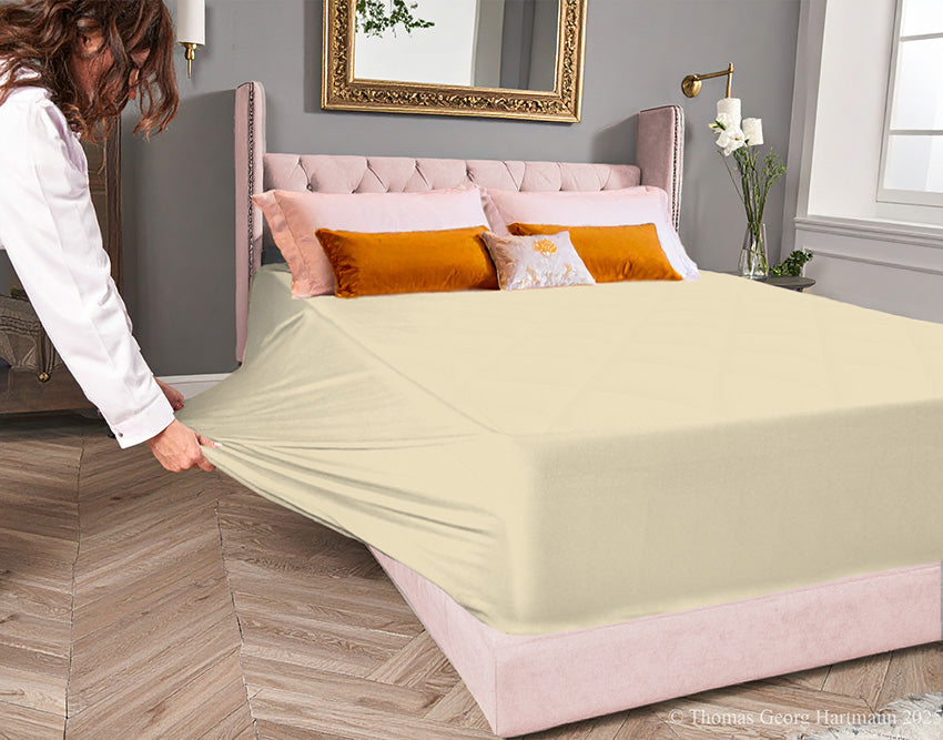 Sand jersey-knit stretch fitted sheet on an extra-deep mattress — pulling the sidewall over a foot outward to demonstrate the abundant stretch, making sheet changing effortless even for those with limited strength or mobility.