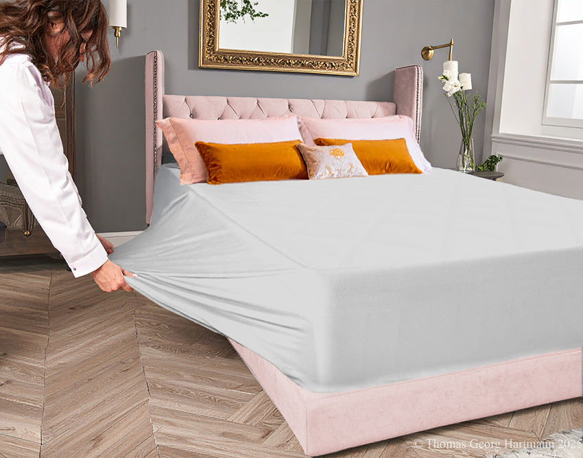 Silver jersey-knit stretch fitted sheet on an extra-deep mattress — pulling the sidewall over a foot outward to demonstrate the abundant stretch, making sheet changing effortless even for those with limited strength or mobility.