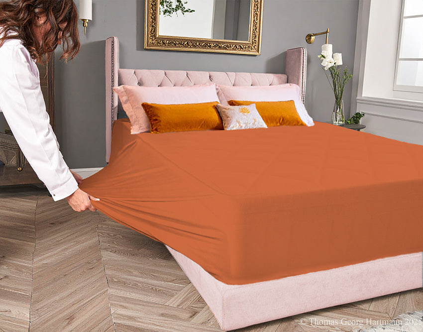 Terracotta jersey-knit stretch fitted sheet on an extra-deep mattress — pulling the sidewall over a foot outward to demonstrate the abundant stretch, making sheet changing effortless even for those with limited strength or mobility.
