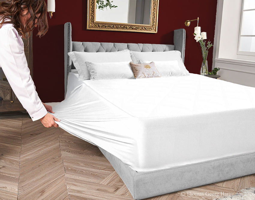 White jersey-knit stretch fitted sheet on an extra-deep mattress — pulling the sidewall over a foot outward to demonstrate the abundant stretch, making sheet changing effortless even for those with limited strength or mobility.